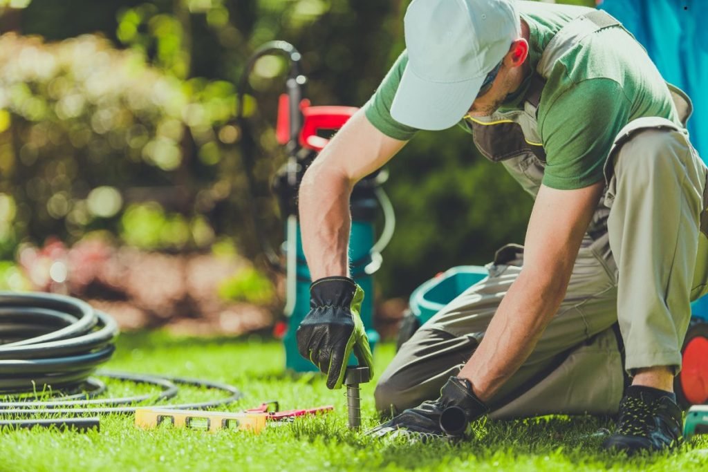 A gardener in gloves installs equipment on the grass, surrounded by tools and hoses, focused on landscaping work