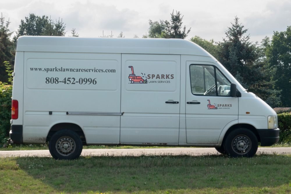 A white van parked on grass, displaying a sign on its side, surrounded by greenery.